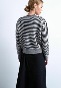 Femme aux cheveux châtain clair attachés en chignon, portant un pull rayé noir et blanc et une longue jupe noire, vue de dos sur un fond clair uni.