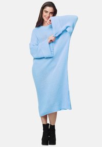 Abito maglione sovradimensionato di colore azzurro chiaro con maniche larghe e spacchi laterali, indossato sopra stivali neri sopra la caviglia. Tessuto texturizzato, vestibilità comoda.