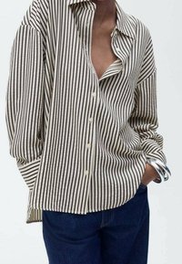 Chemise à rayures beige et noire avec boutons, manches longues, coupe décontractée et col en V profond. Portée avec un jean bleu et un bracelet argenté.