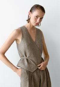 Le modèle pose dans un haut en lin brun avec une taille peplum. Elle a les cheveux mouillés et porte des boucles d'oreilles, devant un fond blanc uni.