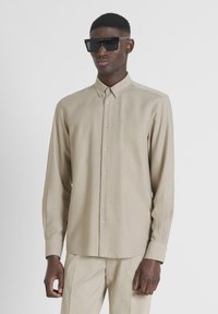 Camisa de manga larga beige hecha de tejido ligero, con cuello abotonado, botones frontales y puños ajustados. Llevada con gafas de sol negras.
