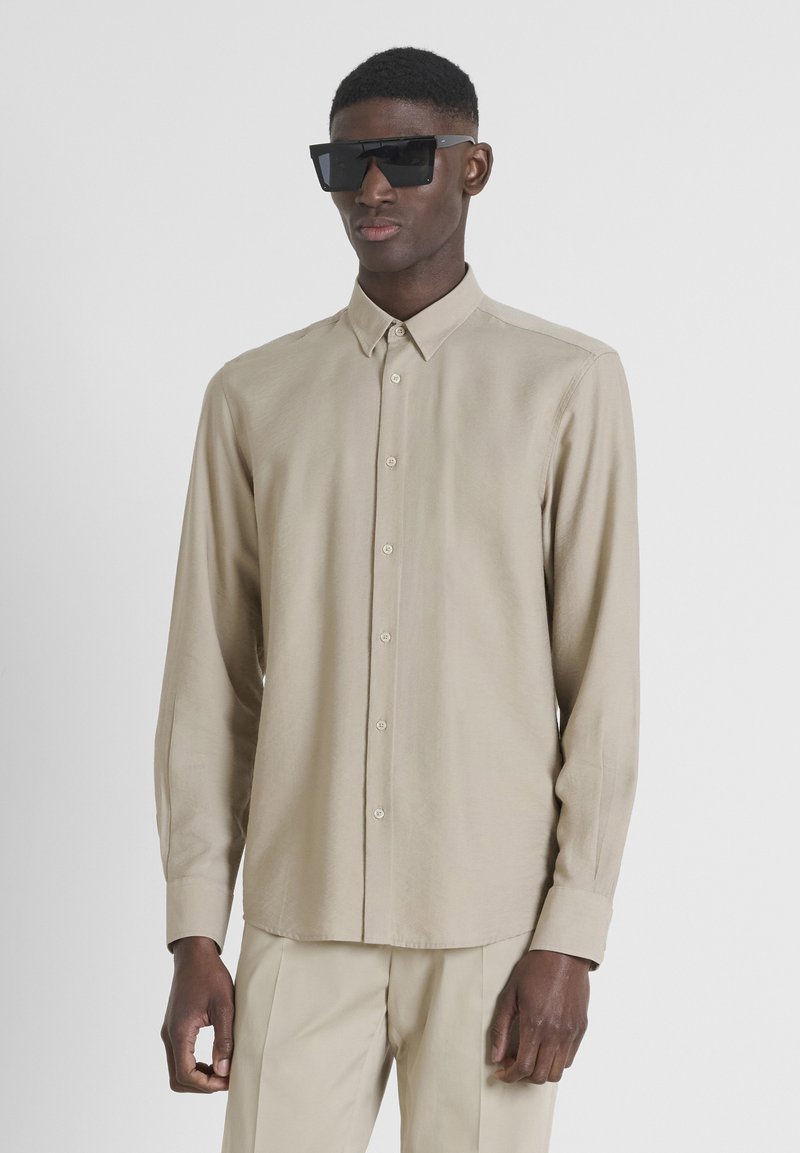 Camisa de manga larga beige hecha de tejido ligero, con cuello abotonado, botones frontales y puños ajustados. Llevada con gafas de sol negras.