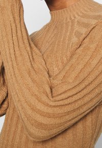 Maglione lavorato a maglia in beige chiaro, caratterizzato da una trama a coste e maniche allungate. Ingrandimento che mostra la texture e i dettagli ondulati sul tessuto.