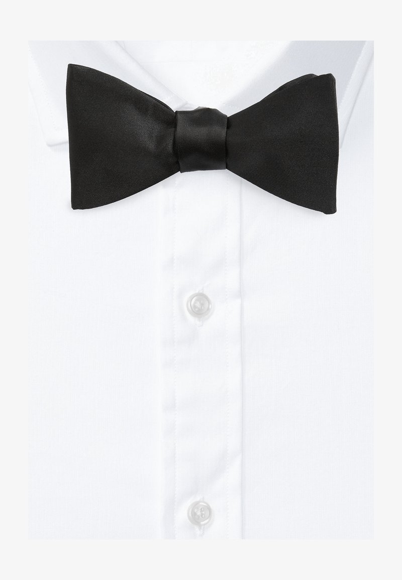 Papillon in raso nero con texture liscia, posizionato al centro di una camicia bianca con colletto a punta e bottoni trasparenti.