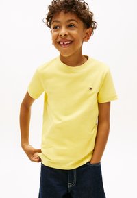 Χαμογελαστός νεαρός αγόρι με σγουρά μαλλιά, φορώντας κίτρινη κοντομάνικη μπλούζα και σκούρο τζιν σορτς, στέκεται με το ένα χέρι στην τσέπη και το άλλο τραβώντας τη μπλούζα.