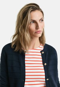 Navy Strickjacke mit strukturierten Streifen und goldenen Knöpfen, über einem weißen Hemd mit roten horizontalen Streifen getragen.