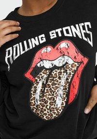 Melna džemperis ar nolietotu grafiku, kas attēlo sarkanas lūpas ar leoparda raksta mēli un tekstu "ROLLING STONES" treknajās baltajās burtos.