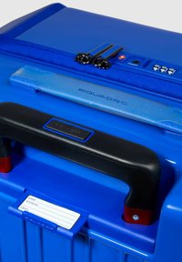 Valigia rigida blu con manico nero, dettagli rossi, lucchetto a combinazione trasparente e porta etichetta per nome e indirizzo.