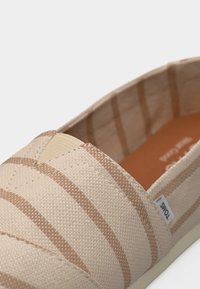 Μπεζ καμβαδένιο παπούτσι τύπου slip-on με καφέ οριζόντιες ρίγες και μια μικρή υφασμάτινη ετικέτα με την ένδειξη TOMS στο πλευρό.