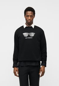 Άνδρας φοράει μαύρο φούτερ με λευκό πουκάμισο με γιακά από κάτω, με γραφικό σχέδιο λευκών ριγέ γυαλιών ηλίου και κείμενο "KARL LAGERFELD".