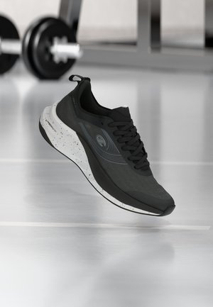 Chaussure de sport noire avec semelle blanche tachetée flottant à l’intérieur près d’haltères flous et d’équipements de gym en arrière-plan.