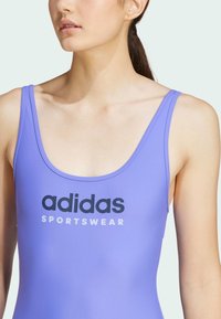 Lila baddräkt i ett stycke med breda axelremmar och rundad halsringning, tillverkad av ett slätt tyg med svart "adidas SPORTSWEAR"-logotyp på framsidan.