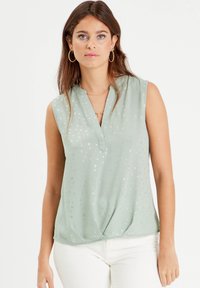Blouse sans manches vert clair avec un décolleté en V et un détail en torsion à l'avant. Présente un motif de petits points argentés éparpillés. Tissu lisse.