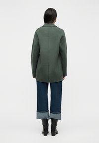 Γυναίκα με μακριά σκούρα μαλλιά, που φοράει πράσινο παλτό, σκούρα μπλε τζιν με γυρισμένα μανίκια και μαύρα μποτάκια με τακούνι, στέκεται με την πλάτη γυρισμένη σε λευκό φόντο.