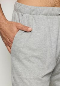 Pantaloni de trening gri deschis, dintr-un material texturat, cu un elastic la talie. Prezintă o buzunar lateral și accente de cusătură vizibile.