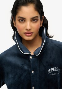Veste en velours côtelé bleu marine avec un col haut et des boutons-pression en métal argenté. Présente des bordures blanches et un logo "SUPERDRY" brodé sur la poitrine.