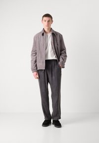 Veste en velours côtelé gris avec boutons-pression, chemise blanche et pantalon noir foncé ample. Des chaussures noires à enfiler complètent la tenue.