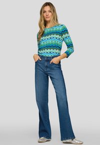 Langärmliges Oberteil mit blau-grünem Zickzackmuster, kombiniert mit hoch taillierter blauer Jeans und silbernen flachen Schuhen.