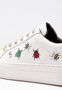Bílé kožené tenisky s vyšitými vzory hmyzu: zelený brouk, slunéčko, dvě včely a moucha. Obsahují zlaté tkaničky a gumovou podrážku.