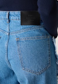 Hochgeschnittene blaue Denim-Jeans mit strukturiertem Oberflächen, fransigem Saum und einem schwarzen Lederetikett mit dem eingeprägten Schriftzug "MADS NØRGAARD COPENHAGEN".