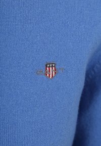 Blauer Strickstoff mit einem kleinen gestickten Wappenlogo, das Sterne und Streifen sowie das Wort "GANT" in grauem Faden zeigt.