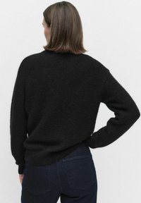 Kvinna med axellångt brunt hår som bär en svart fluffig tröja och mörkblå jeans, sedd bakifrån mot en enkel bakgrund.