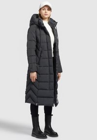 Lungo cappotto imbottito nero con cappuccio, chiusura con cerniera frontale, motivo a rombi diagonali e tasche laterali. Indossato con pantaloni neri e stivali neri.