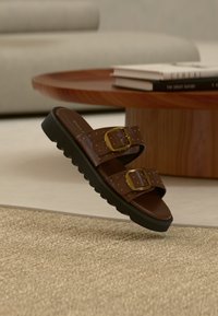 Sandales en cuir marron avec des brides à double boucle, des accents dorés et une semelle texturée noire. Flottant au-dessus d'une surface recouverte de moquette beige.