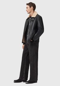 Sort læderjakke med en tanfarvet shearling-krave, knaplukning foran, sidelommer, kombineret med løstsiddende mørke jeans og sorte sko.