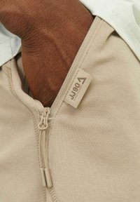 Vêtement beige à fermeture éclair avec tissu texturé, arborant une étiquette avec le logo "JJ 190" et un design bicolore. Gros plan sur la fermeture éclair et le tissu.