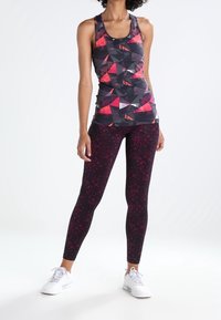 Czarny top na ramiączkach z czerwonym geometrycznym wzorem, zestawiony z ciemnymi, wzorzystymi legginsami. Modelka ma na sobie białe buty sportowe.