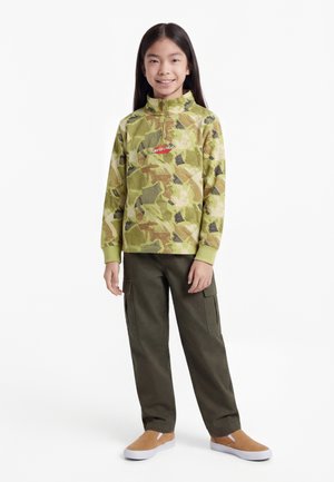 Kind staat en lacht, draagt een groene camouflage kwart-rits trui, donkere cargobroek en beige instapschoenen tegen een witte achtergrond.