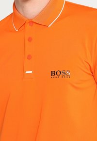 Polo orange avec un col texturé, trois boutons et une bordure blanche. Présente le logo "BOSS" en noir de manière bien visible.