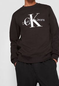 Černá mikina z bavlněné směsi s velkým bílým logem "Calvin Klein Jeans" v moderním písmu. Žebrovaný výstřih a manžety.