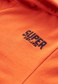 Narančasti materijal s crnim izvezenim logotipom "SUPER DRY.JPN", prikazujući teksturu i detalje šavova.