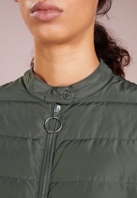 Tmavě zelená péřová bunda s vysokým límcem, předním zipem a kulatým táhlem zipu. Obsahuje prošívané části a zapínání na patentku.