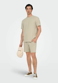 Beige tekstureret kortærmet skjorte og matchende shorts, parret med hvide sneakers og en lys farvet cap holdt i den ene hånd.