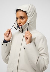 Veste imperméable beige clair avec un col montant, une capuche réglable, une fermeture éclair noire et des poignets à pression ; présente une texture lisse et mate.