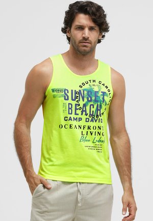 Man with curly dark hair wearing a bright yellow tank top with blue and black beach-themed text and light beige pants.