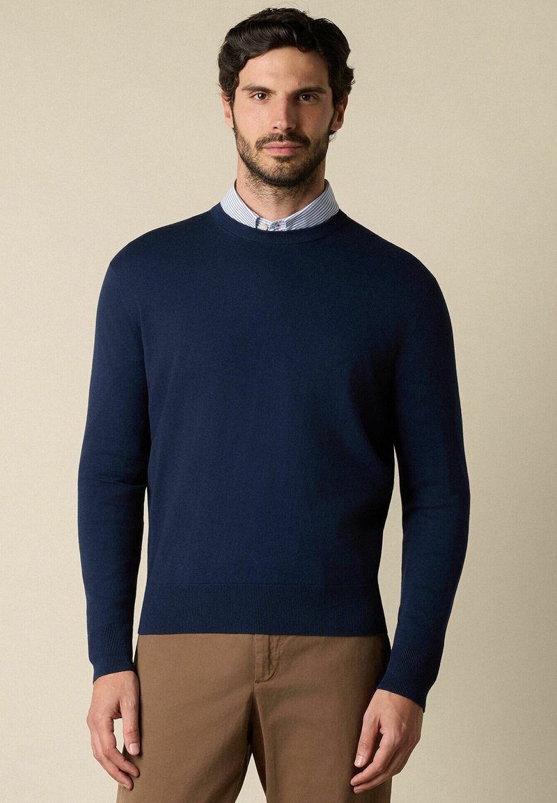 Marineblauer Rundhals-Pullover aus einem glatten Strickstoff, mit langen Ärmeln und einem gerippten Saum, übereinander getragen mit einem hellblau karierten Hemd.