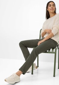 Olivgrüne Baumwollhosen mit schlanker Passform, kombiniert mit einem cremefarbenen Pullover und beigen Sneakern, sitzend auf einem grünen Stuhl vor einem minimalistischen Hintergrund.