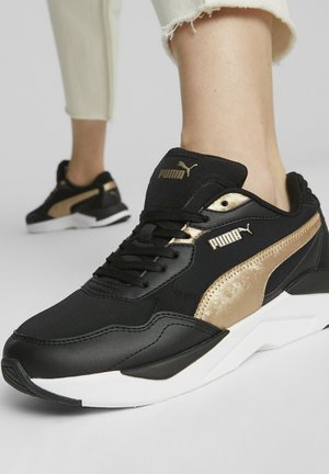 Sorte og gyldne Puma sneakers båret med beige korte bukser, der viser detaljerede side- og frontvisninger med hvide såler.
