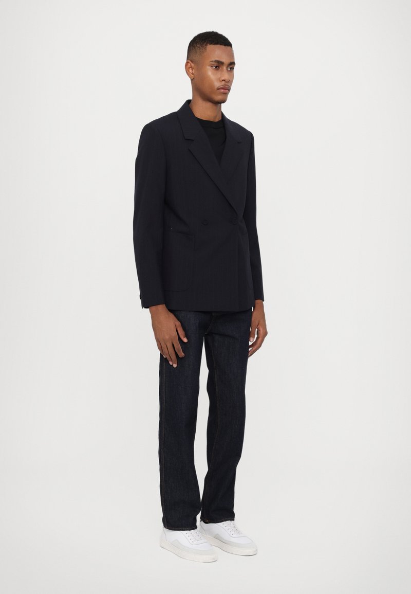 Marineblauer, doppelt geknöpfter Blazer mit Reverskragen und frontalen Taschen, kombiniert mit dunklen Jeans und weißen Sneakers. Glattes Material und taillierte Passform.