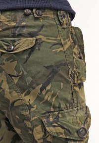 Kamouflage cargo-byxor med flera knapplösa fickor, bärs med en mörk stickad topp som visar midja och övre lår.