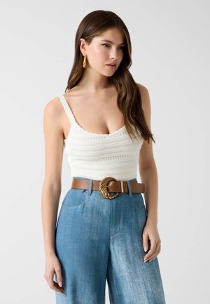 Mujer con cabello largo y castaño que lleva una camiseta de punto blanca sin mangas, vaqueros azules de pierna ancha y cintura alta, y un cinturón marrón con hebilla dorada.