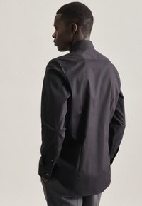 Chemise noire à manches longues et à col classique, texture lisse, coupe ajustée et poignets à boutons, portée avec un pantalon gris.