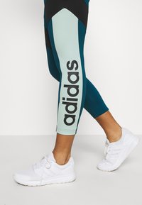 Legginsy mają design w kolorze blokowym w odcieniach teal i miętowej zieleni, z dużym czarnym logo "adidas" wzdłuż boku; zestawione z białymi butami sportowymi.