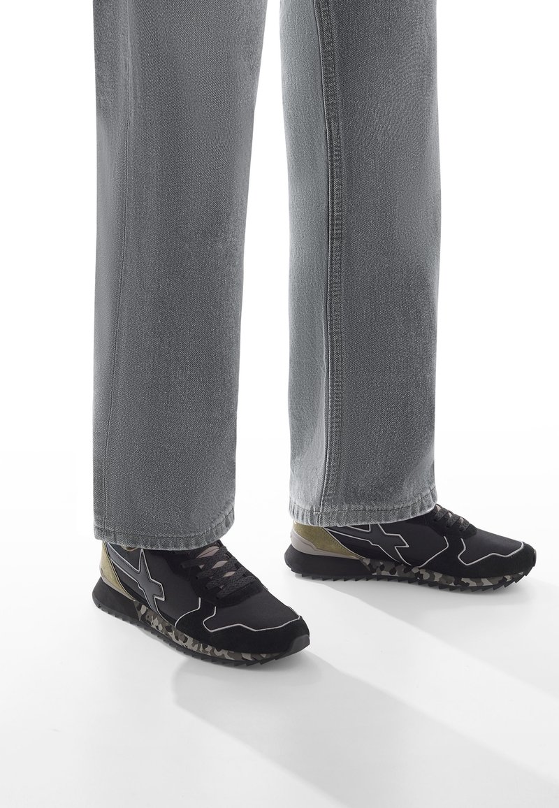 Jambes inférieures portant un jean gris et des baskets noires avec des accents beiges et des semelles à motifs, debout sur une surface blanche.