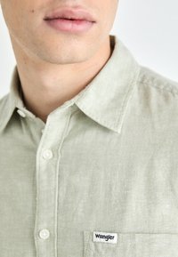 Light green linen shirt with a collared neck, button closure, and a chest pocket featuring a black "Wrangler" label.