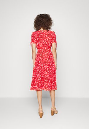 Femme debout, dos tourné, portant une robe midi rouge à fleurs avec des manches bouffantes et des sandales à talons hauts beige sur un fond uni.